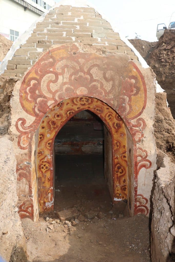 La puerta de la tumba está pintada con diseños botánicos rojos y amarillo-naranjas. A cada lado del interior de la puerta hay un par de hombres con túnicas amarillas.