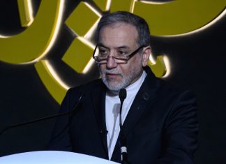 Irán sobre la guerra económica de EEUU: Misma película diferentes matones La fotografía muestra al canciller iraní durante una intervención pública. Aparece vestido con traje oscuro y camisa blanca, detrás de un atril y frente a varios micrófonos, mientras al fondo se observa un elemento gráfico de gran tamaño.