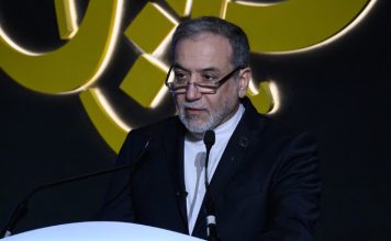 Irán sobre la guerra económica de EEUU: Misma película diferentes matones La fotografía muestra al canciller iraní durante una intervención pública. Aparece vestido con traje oscuro y camisa blanca, detrás de un atril y frente a varios micrófonos, mientras al fondo se observa un elemento gráfico de gran tamaño.