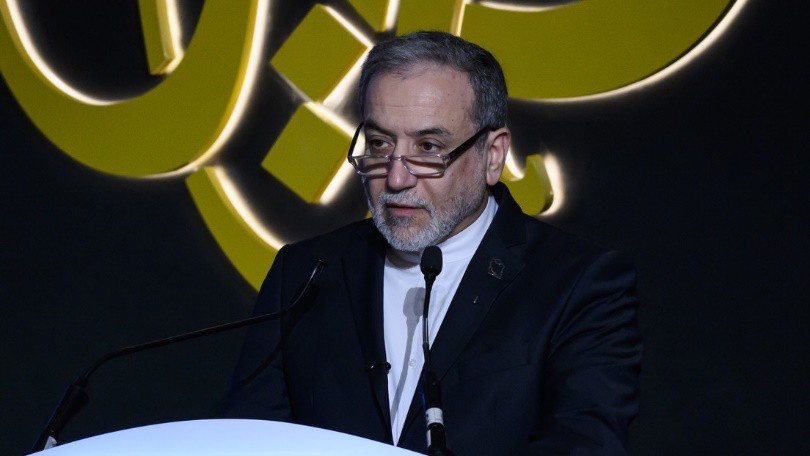 La fotografía muestra al canciller iraní durante una intervención pública. Aparece vestido con traje oscuro y camisa blanca, detrás de un atril y frente a varios micrófonos, mientras al fondo se observa un elemento gráfico de gran tamaño.