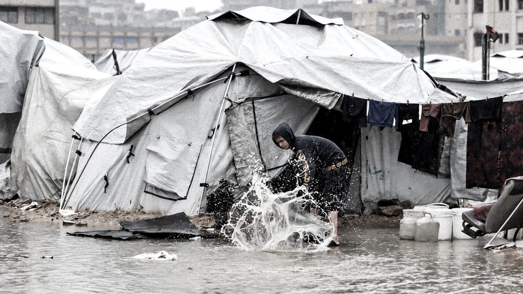 Niño palestino sacando agua de una tienda inundada en Gaza ante la crisis de refugio.