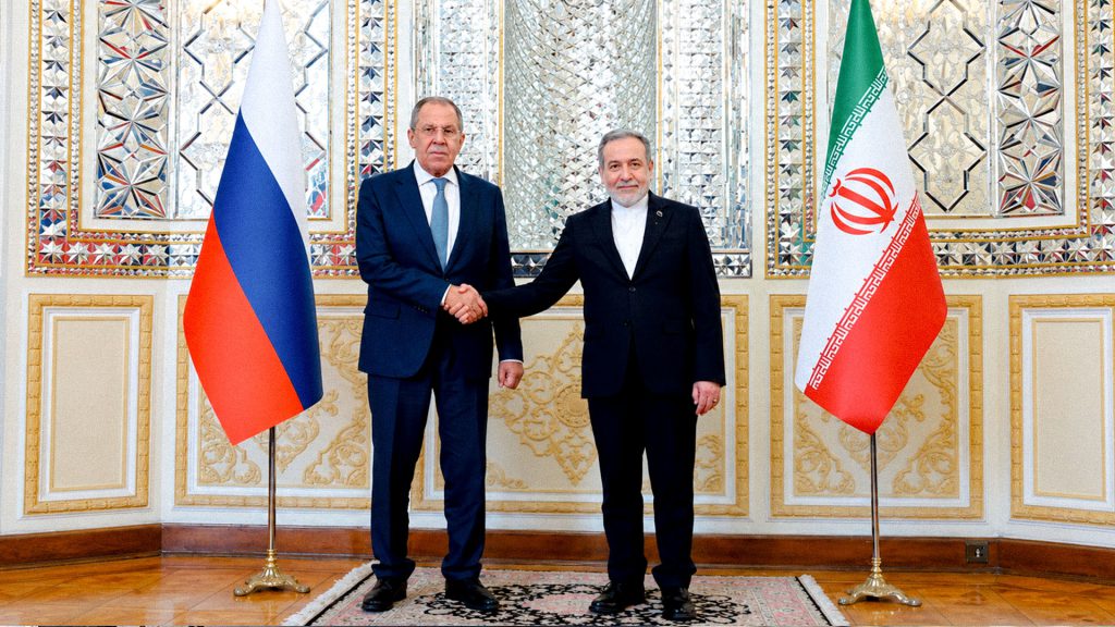 Representantes diplomáticos de Irán y Rusia estrechan sus manos frente a las banderas de ambos países.